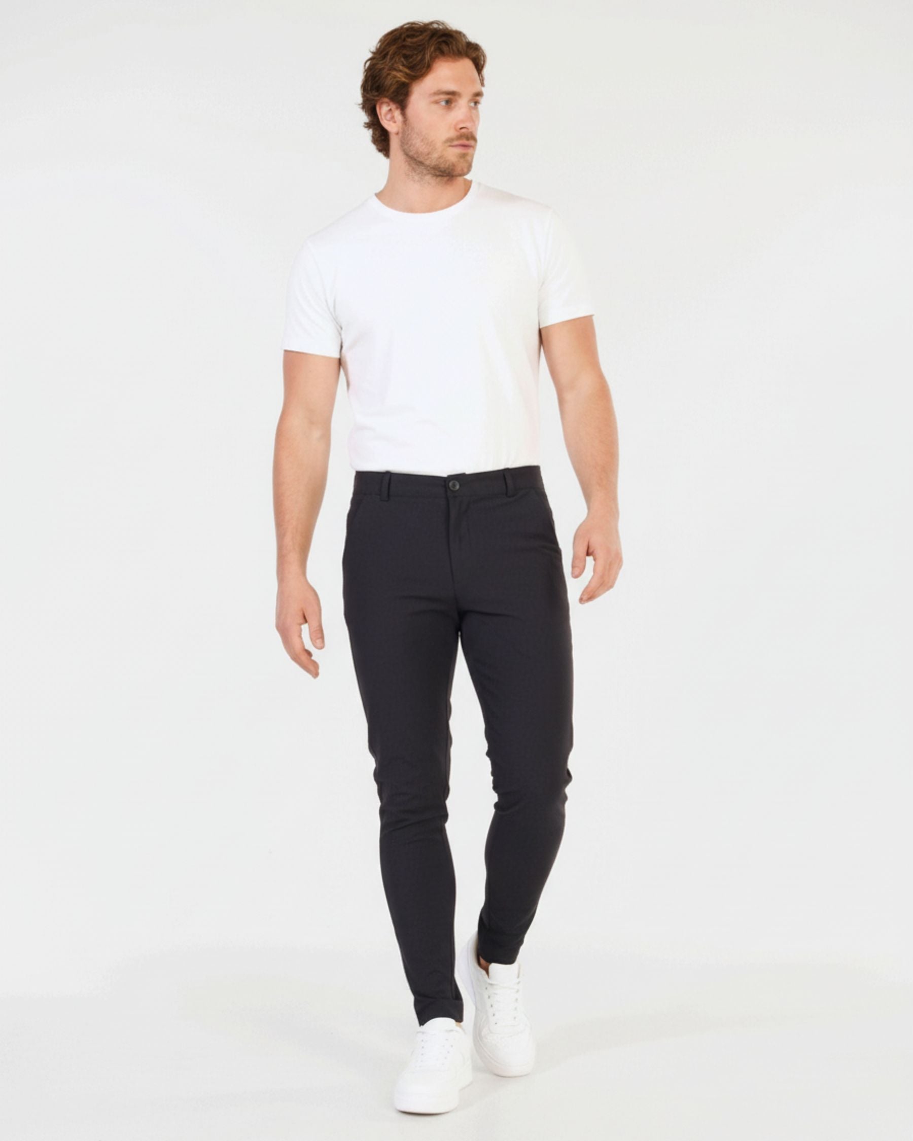 A man modeling a white crew-neck t-shirt and slim-fit black trousers with white sneakers, standing against a solid white studio background.