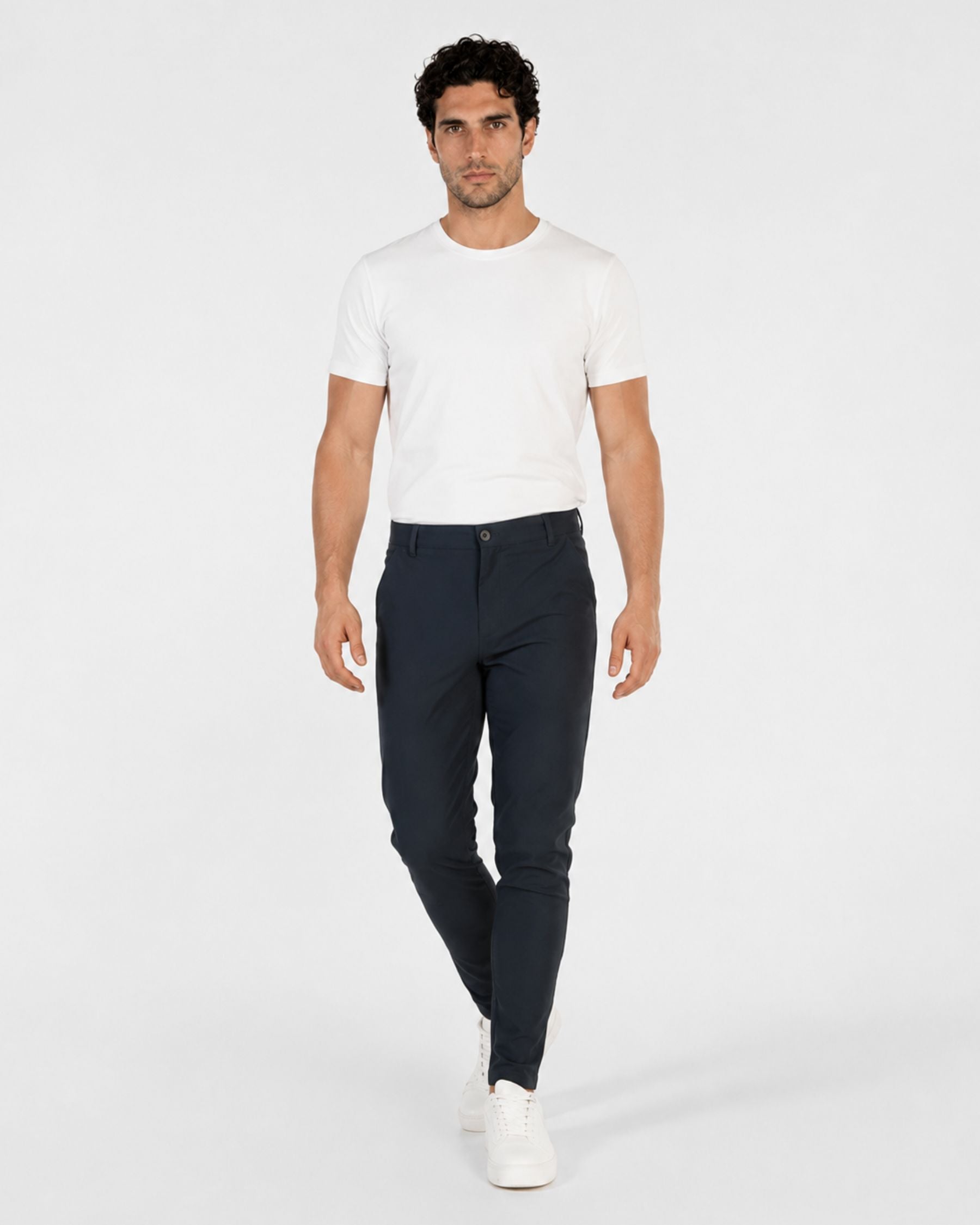 A man modeling a white short-sleeve crewneck t-shirt and dark navy blue slim-fit trousers, paired with white sneakers, against a plain light gray studio background.