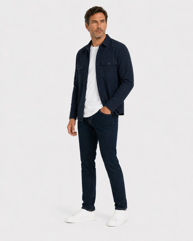 Man wearing a By Alden navy shirt over a white t-shirt, dark jeans, and white sneakers on a light gray background #color_navy