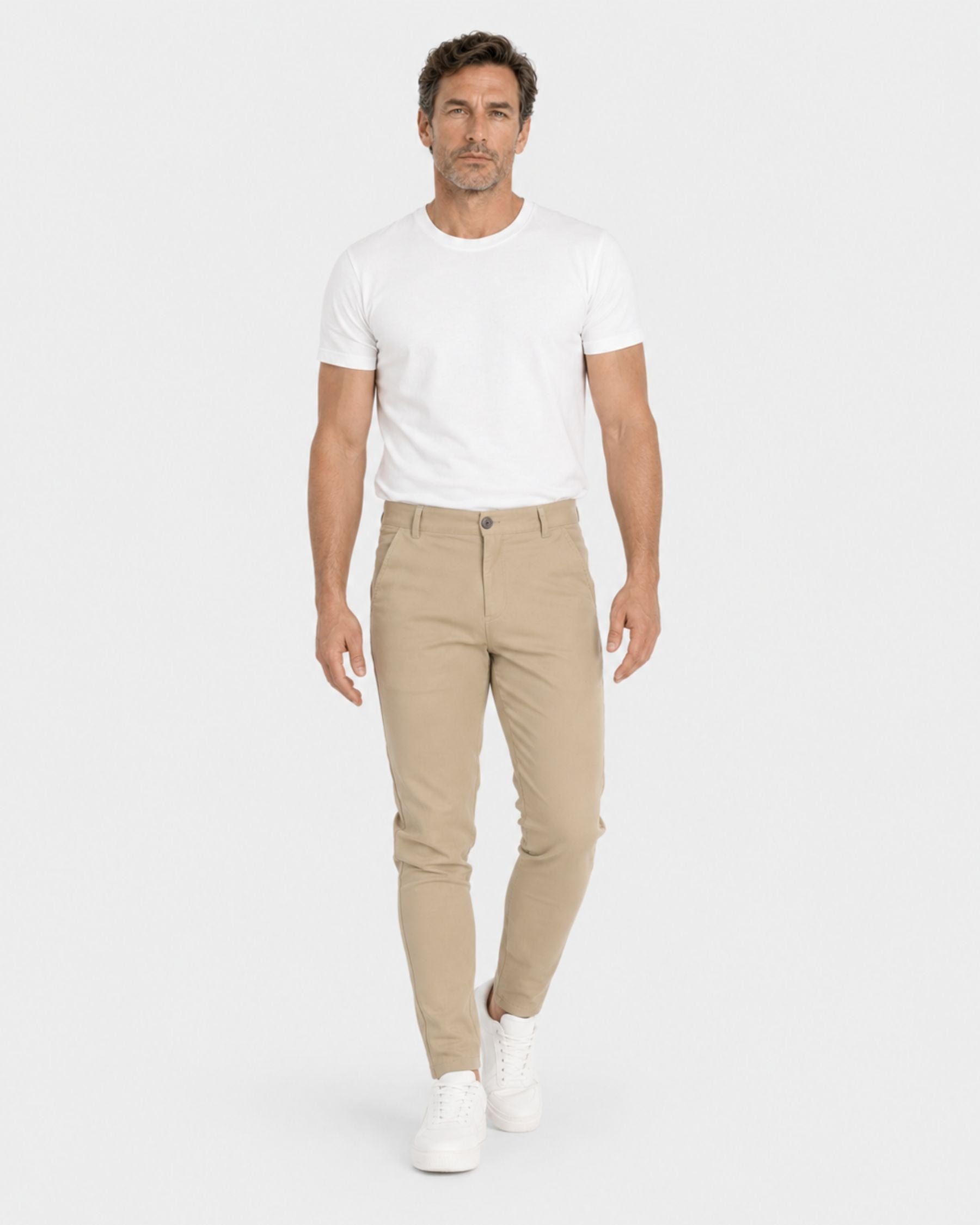 A man modeling slim-fit beige chinos with a white t-shirt and white sneakers, standing against a light gray studio background.