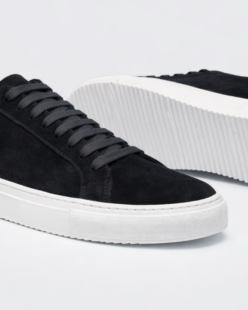 Close-up of minimalist luxury black suede trainers highlighting the textured white rubber sole and premium stitching. #color_black